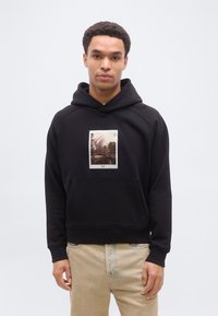 Νέος άνδρας που φοράει μαύρο φούτερ με κουκούλα με εκτύπωση φωτογραφίας ορεινού τοπίου και μπεζ τζιν, στέκεται μπροστά σε απλό ανοιχτό φόντο.