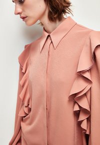 Blusa rosa con un cuello estructurado, que presenta una tela texturizada y llamativas volantes que se extienden desde los hombros. Cierre frontal con botones.