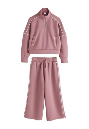 Ljubičasto dvodijelno odijelo s kratkom majicom na zatvarač s bijelim obrubom i pripadajućim širokim hlačama sa bočnim prugama i patentnim zatvaračem u struku.