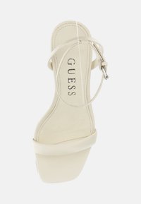 Sandalo con tacco alto beige, con cinturino sottile, punta rotonda e soletta imbottita. Presenta il logo "GUESS" sulla soletta interna e un design con tacco spesso.