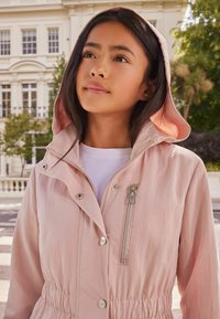 Chaqueta de color rosa claro con capucha, cierres de presión y cremallera, cintura elástica y un bolsillo en el pecho con cremallera, que presenta una tela suave y ligera.
