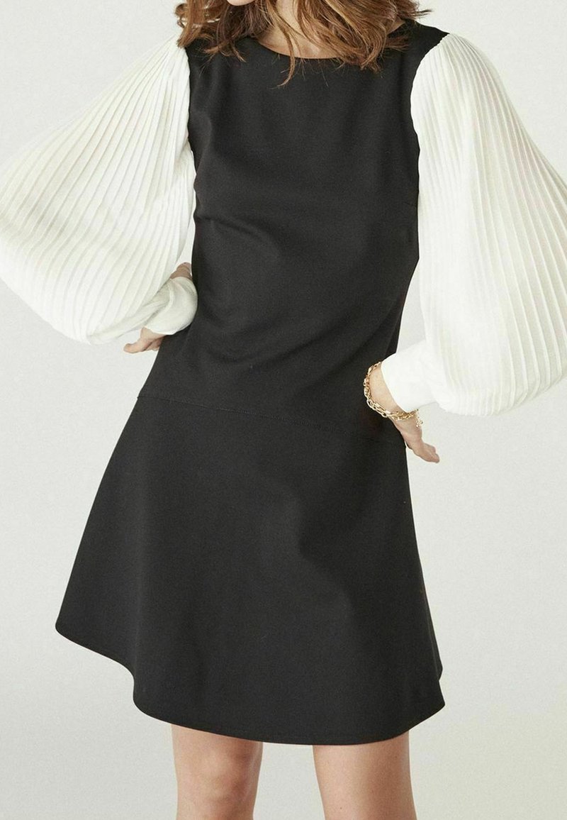 Femme portant une robe noire avec de volumineuses manches longues blanches plissées et un bracelet en or à son poignet droit.