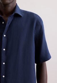 Camicetta blu navy a maniche corte in tessuto leggero, con chiusura frontale a bottoni, colletto a punta e una sottile textura.