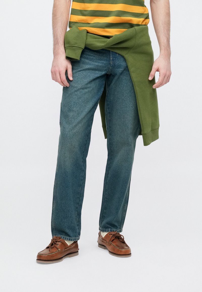 Jeans in denim blu medio, caratterizzati da un taglio dritto e una texture leggermente sbiadita. Abbinati a una felpa verde annodata in vita. Scarpe marroni.