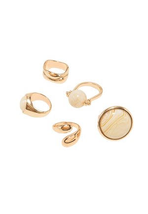 Cinq bagues ton or, dont deux avec des pierres rondes beiges et trois avec des motifs métalliques lisses ou torsadés sur fond blanc.