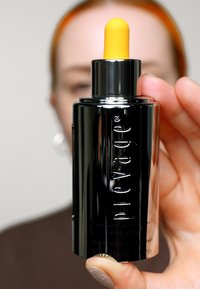 Svart och silver serumflaska med en gul pipett, med präglad text för varumärke och produkt, hålls i en hand mot en suddig bakgrund.