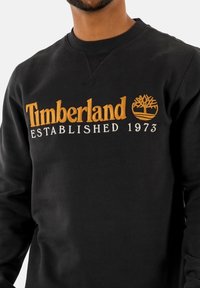 Sweat-shirt noir en mélange de coton, avec logo "Timberland" brodé en orange et un motif d'arbre, col rond et poignets côtelés.