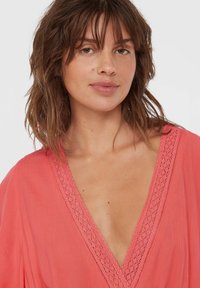 Koraalkleurige blouse met een diepe V-hals, textuurachtige kantdetails langs de halslijn en een vloeiend, lichtgewicht stof. Casual ontwerp.