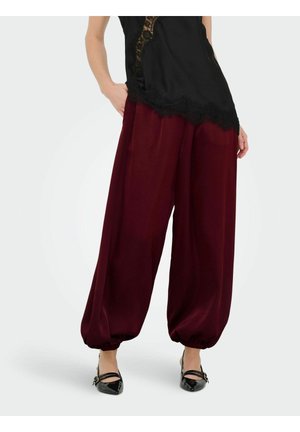 Kobieta w bordowych spodniach o szerokich nogawkach z elastycznymi mankietami, połączonych z czarną koszulką na ramiączkach ozdobioną koronką oraz czarnymi szpilkami z zaostrzonym noskiem.