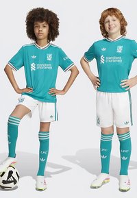 Türkiissinised jalgpalliformid valgete lühikeste pükstega, millel on logo ja brändi detailid. Mängijad kannavad triibulisi sokke ja krampe ning hoiavad musta-valget jalgpallipalli.