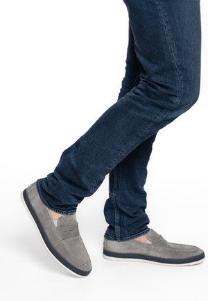 Person trägt dunkelblaue Jeans und graue Wildleder-Slipper, ein Fuß leicht angewinkelt vor weißem Hintergrund.