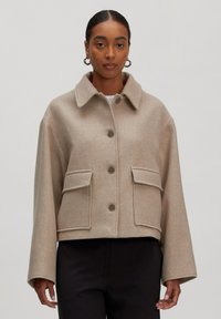 Vrouw in een beige wollen cropped jack met grote klepzakken en knopen, gecombineerd met zwarte broek, op een effen lichte achtergrond.