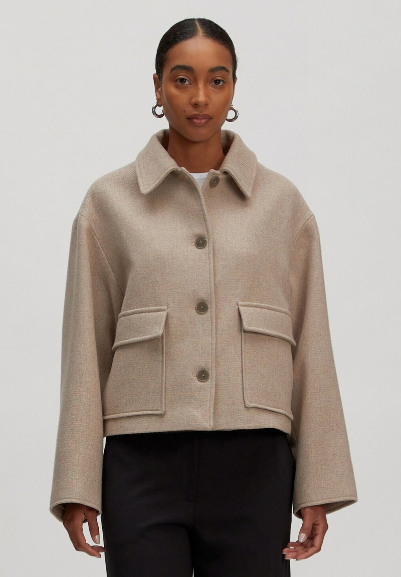 Vrouw in een beige wollen cropped jack met grote klepzakken en knopen, gecombineerd met zwarte broek, op een effen lichte achtergrond.