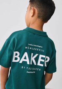 Mladá osoba s krátkymi tmavými vlasmi, oblečená v tmavozelenej košeli Ted Baker, na ktorej je veľký text "BAKER" a menšie logo na chrbte.
