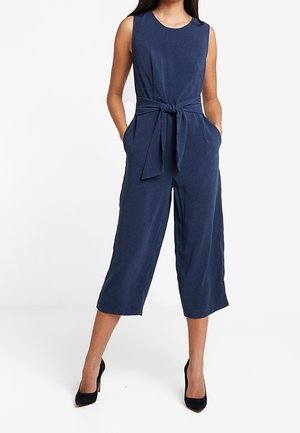 Ærmeløs marineblå jumpsuit med vide, korte ben, bindebånd i taljen, sidelommer, båret med sorte højhælede sko.