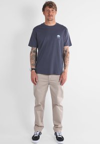 T-shirt en coton gris foncé avec un petit graphisme bleu sur la poitrine, associé à un pantalon beige et des baskets noires avec des lacets blancs.