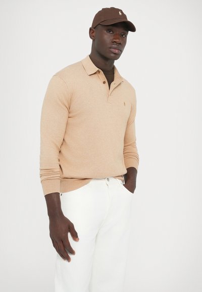 Bézs színű hosszú ujjú póló gallérral és bordás mandzsettával, fehér nadrággal párosítva. A megjelenést egy barna sapka egészíti ki, hímzett logóval.