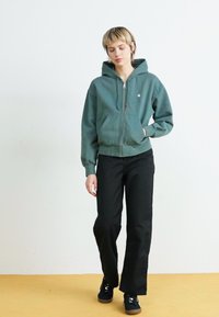 Hooded zip-up sweatshirt in dark green cotton blend, featuring side pockets. Paired with black wide-leg pants and black sneakers.