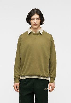 Tânăr cu păr închis de lungime medie, purtând un hanorac verde măsliniu peste o cămașă bej cu guler și pantaloni verde închis.