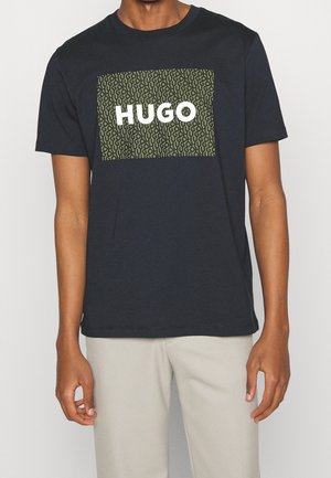 Navy cotton t-shirt featuring a rectangular olive green graphic with repeated "HUGO" text in white. Short sleeves and a classic fit.