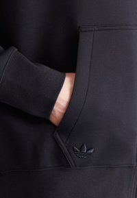 Svart huvtröja med känguruficka, med ribbade ärmslut. Strukturerad tyg med en präglad logotyp på fickan. Slät, modern design.