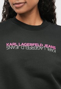 Černá mikina se žebrovaným výstřihem, na které je grafický potisk "KARL LAGERFELD JEANS" v růžové a bílé barvě. Hladký materiál.