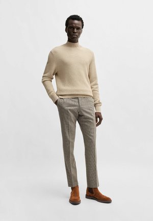 Pull beige texturé avec col haut, associé à un pantalon à carreaux marron clair et des bottines en daim marron.