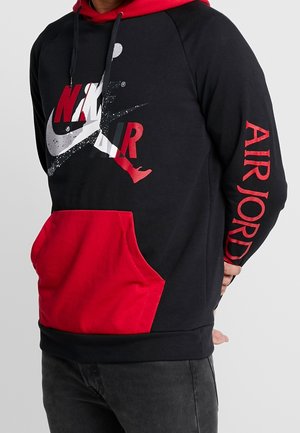Personne portant un sweat à capuche Nike Air Jordan noir et rouge avec un grand logo à l'avant et le texte rouge "AIR JORDAN" sur la manche droite.