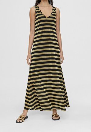 Vrouw die een mouwloze maxi-jurk draagt met zwarte en gouden horizontale strepen en platte sandalen met een luipaardprint band.