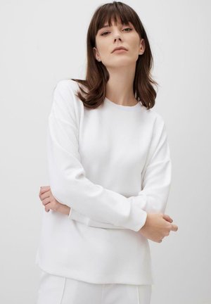 Felpa bianca a maniche lunghe con scollatura a girocollo, texture liscia, vestibilità rilassata e orlo leggermente curvo, indossata con le braccia incrociate.