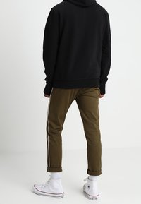 Felpa nera con cappuccio, pantaloni verde oliva con strisce laterali bianche e sneakers bianche alte. Presenta polsini elasticizzati e una vestibilità comoda.