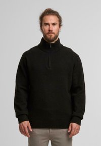 Zwarte wollen pullover met een hoge, geribbelde kraag en een rits tot halverwege. Heeft lange mouwen en een gebreide textuur.