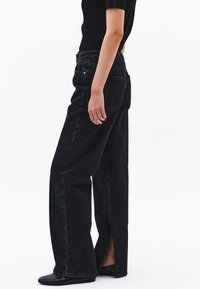 Jeans de denim negro de cintura alta con un diseño de pierna ancha, que presentan aberturas en el dobladillo y bolsillos traseros tradicionales. Tejido texturizado visible.