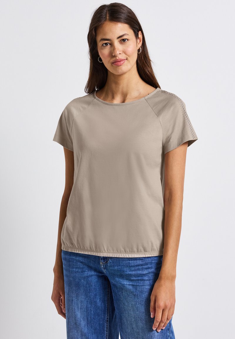 Haut beige clair à manches courtes avec un col rond, accents texturés sur les épaules et ourlet ajustable. Assorti à un jean en denim bleu.