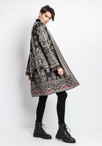 Strikket poncho med indviklede sort-hvide mønstre, røde accenter, oversized form og kontrasterende kant. Båret over sorte leggings og støvler.