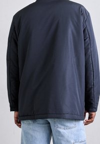 Marinblå vadderad jacka med en slät textur, med rund hals och långa ärmar, kombinerad med ljusblå jeans.