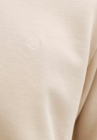 Tissu beige avec une texture fine en maille et un petit logo géométrique blanc brodé discret sur la partie supérieure de la poitrine.