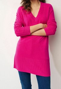 Pull en tricot rose avec un col en V, des poignets côtelés et un ourlet côtelé plus long. Texture douce et coupe décontractée, associé à un jean foncé.