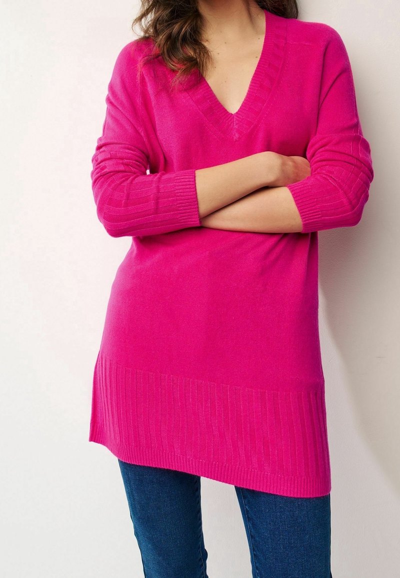 Pull en tricot rose avec un col en V, des poignets côtelés et un ourlet côtelé plus long. Texture douce et coupe décontractée, associé à un jean foncé.
