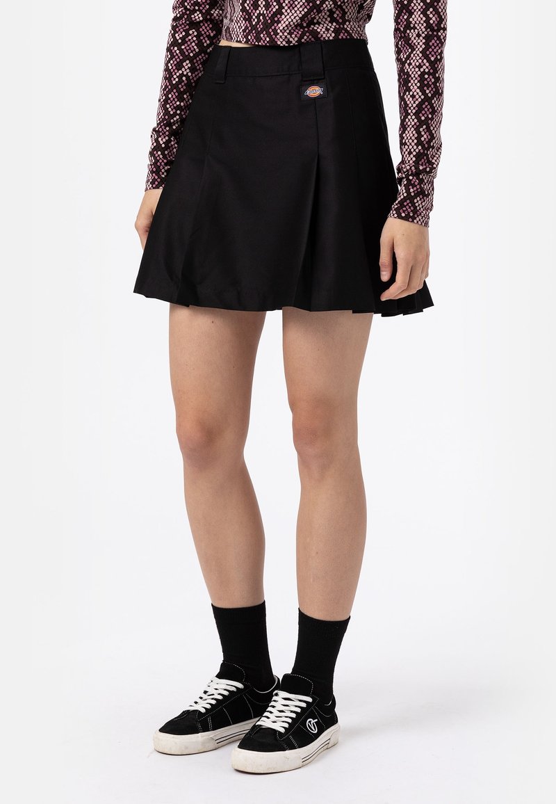 Femme portant une jupe plissée noire, des chaussettes noires, des baskets noires et blanches, et un haut à manches longues à motifs, debout devant un fond blanc.