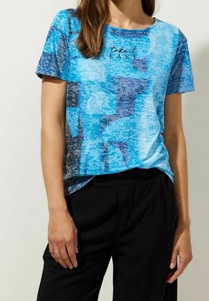 Kobieta ubrana w krótką niebieską koszulkę z nadrukiem „take it EASY” i czarne spodnie, stojąca na jednolitym tle.