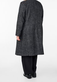 Dlouhý šedý kabát s kostkovaným vzorem, rovného střihu, s dlouhými rukávy a límcem. Spárován s černými kalhotami a lesklými černými botami.