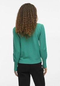 Grüner Pullover mit einem strukturierten Fischgrätmuster, geripptem Saum und Bündchen sowie puffigen Schultern. Kombiniert mit schwarzen Hosen.