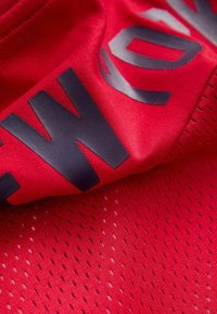 Czerwony sportowy materiał z dużymi czarnymi literami nadrukowanymi na gładkich i siateczkowatych sekcjach, pokazujący zbliżenia detali materiału i wzoru.