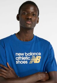 Blå bomulds-T-shirt med grafik i gult og hvidt tekst, der siger "new balance athletic shoes" sammen med New Balance-logoet.