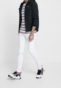 Veste noire à capuche, t-shirt rayé, jean blanc slim et baskets noir/blanc. Tatouages notables sur le bras et logo sur la veste.