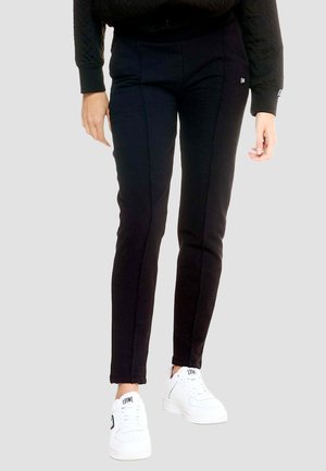 Personne portant un pantalon slim noir et des baskets blanches avec la marque "LEONE", debout devant un fond clair uni.