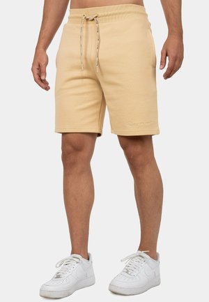 Man draagt beige shorts met trekkoord en witte sneakers, staat met ontspannen armen langs het lichaam tegen een effen achtergrond.