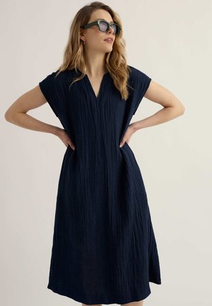 Žena v tmavomodrých midi šatách s krátkym rukávom a textúrou, so slnečnými okuliarmi, stojaca s rukami v bok na jednoduchom pozadí.