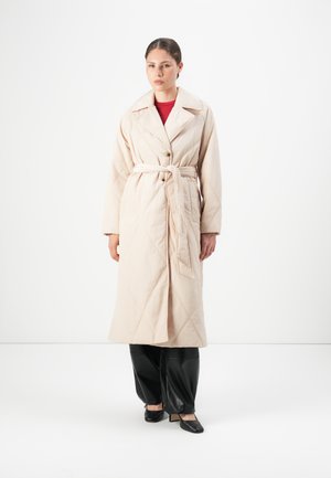 Femme portant un long manteau matelassé beige noué à la taille, un pantalon noir et des chaussures à talons carrés noires, se tenant sur un fond blanc.